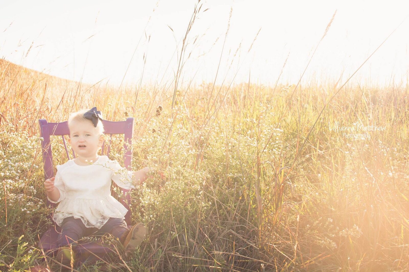 Baby girl seated solo on a purple chair prop in hazy golden light