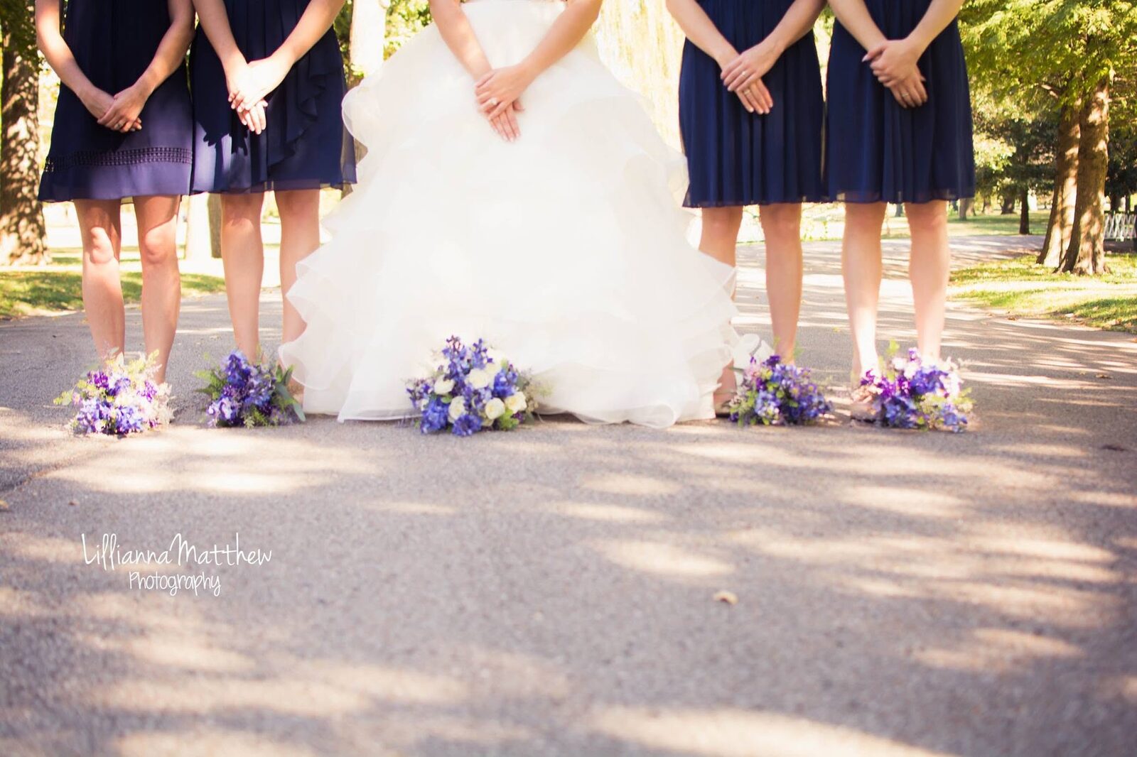 Bride and bridesmaids in navy dresses holding purple and white bouquets, focus on dress and legs