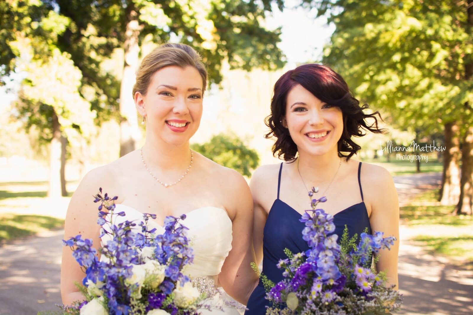 Bride and bridesmaid smiling close together under trees dappled with sunlight