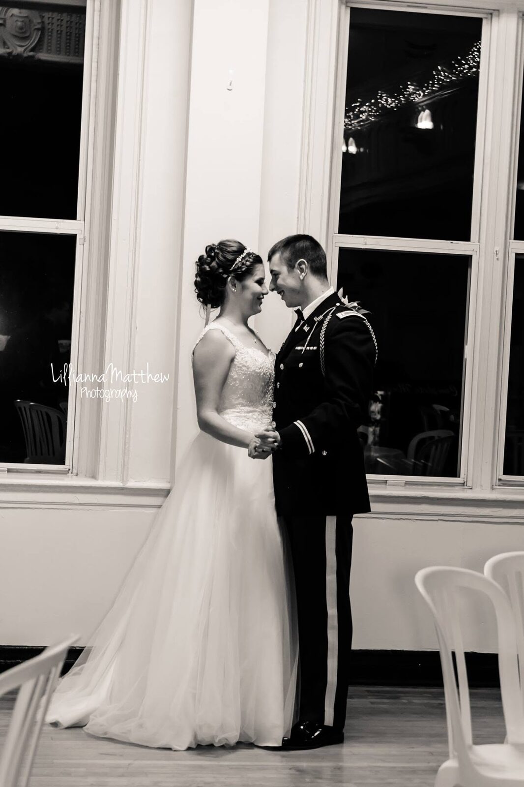 Black and white photo of bride and groom in military dress uniform slow dancing by tall windows