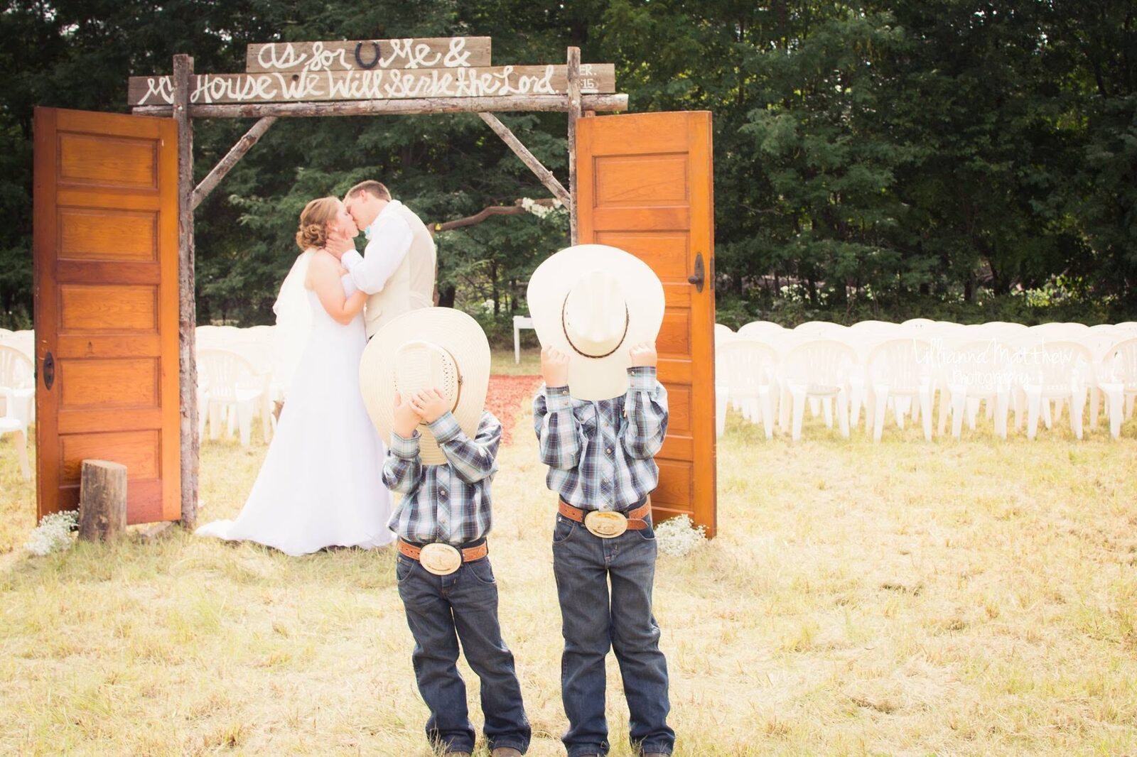 Bride and groom kissing under a rustic wooden arch with two young ring bearers in cowboy hats in the foreground
