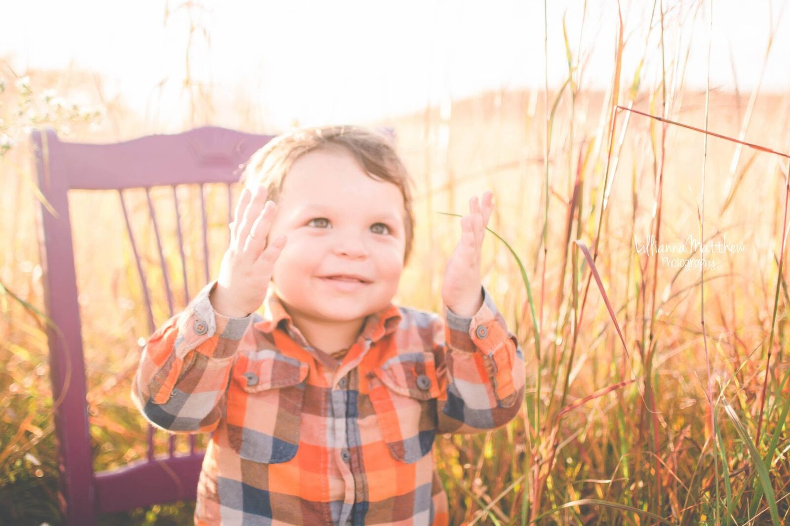 Toddler boy in orange and blue plaid, hands raised, laughing joyfully on a purple chair prop