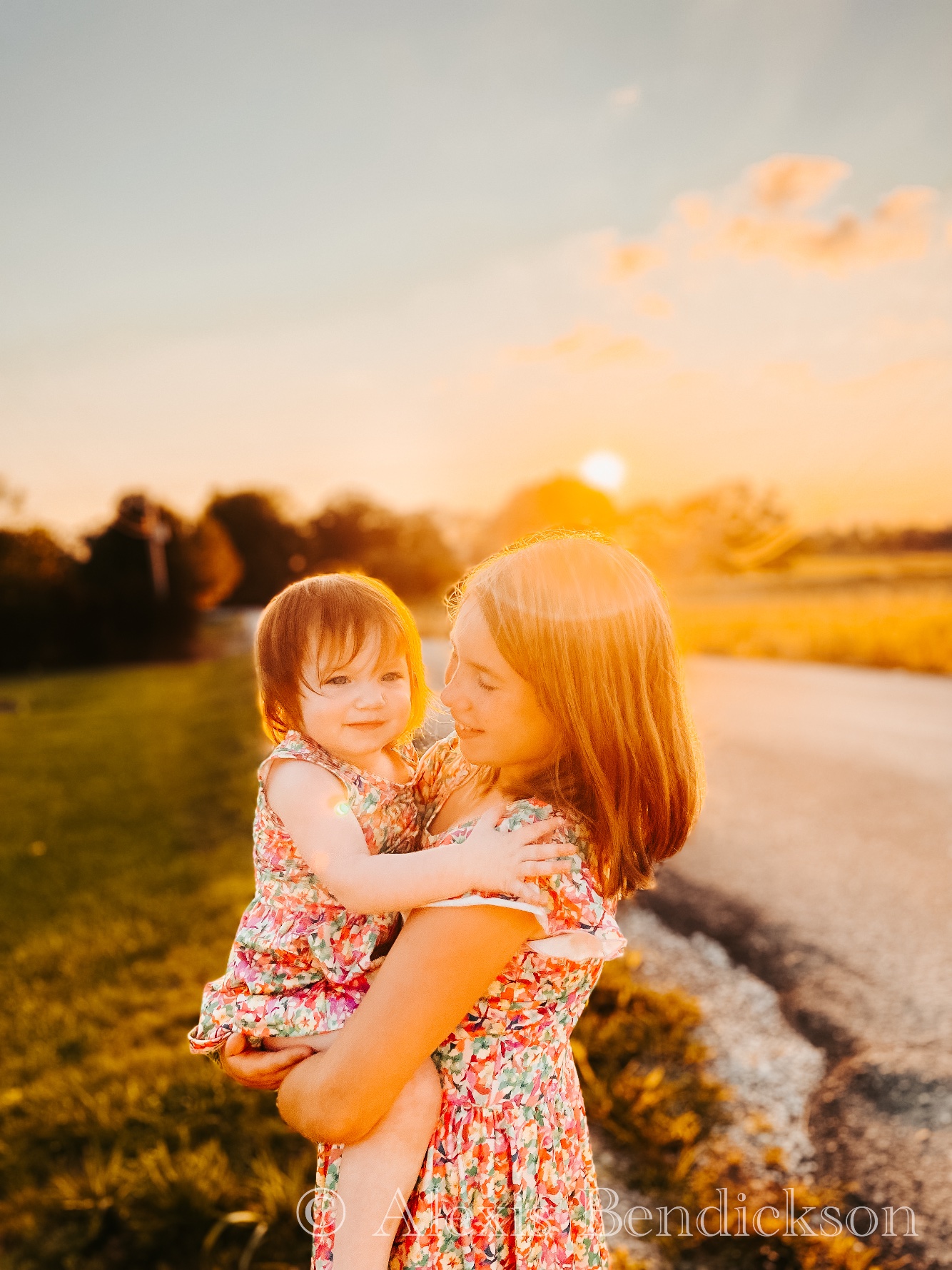 Arianna holding baby Emme at sunset, both in floral dresses
