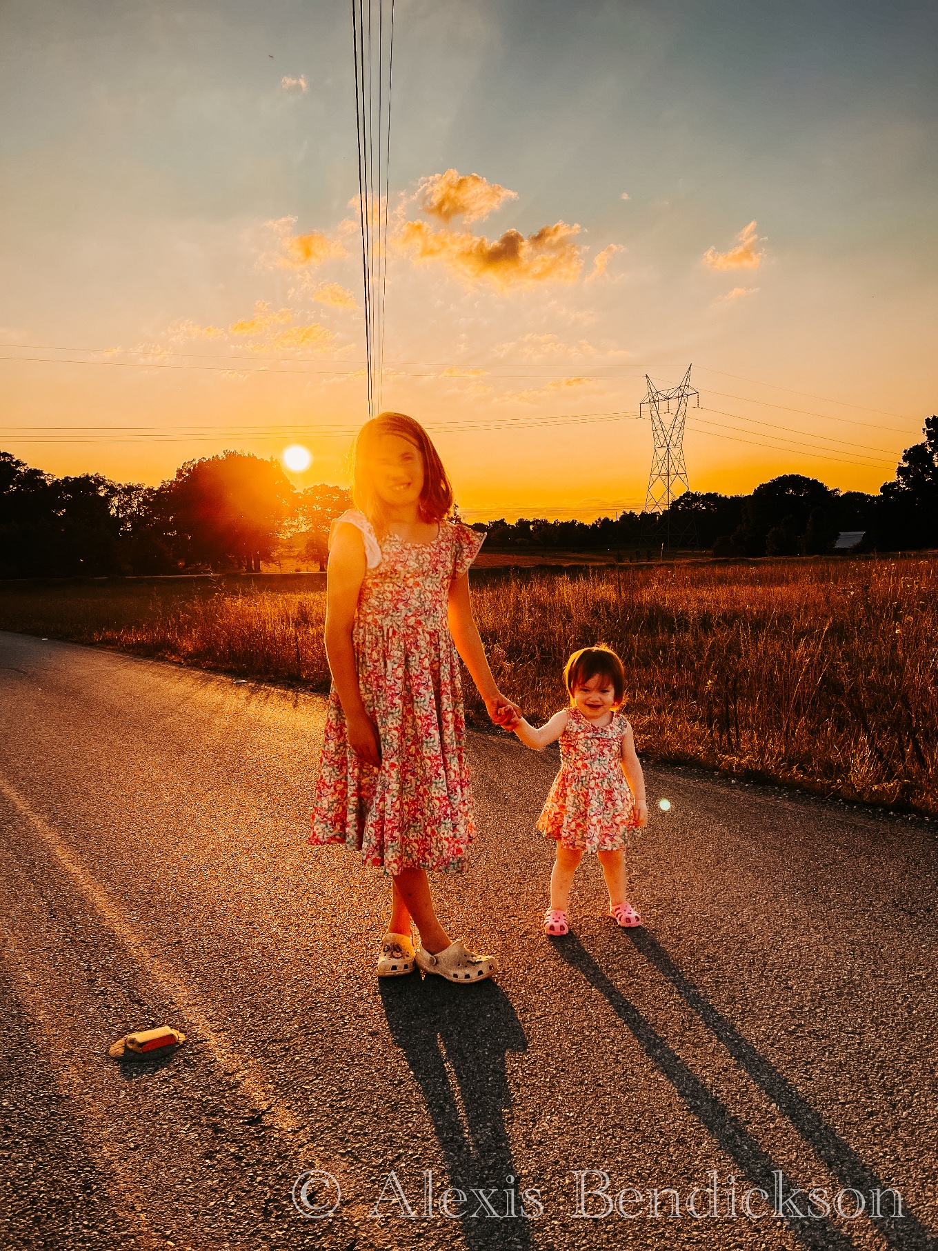Arianna and Emme holding hands on a road at sunset, sun flare