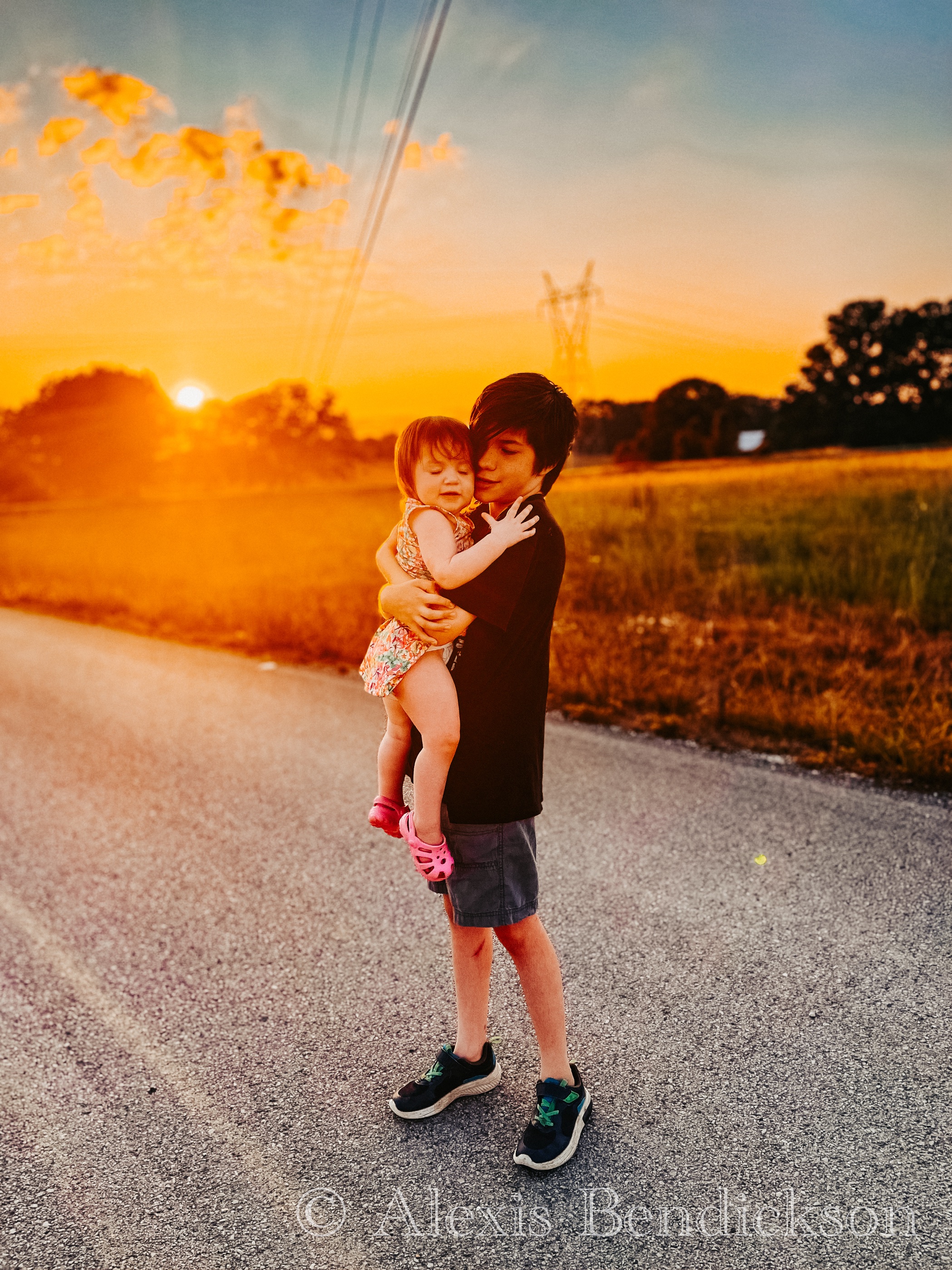 Noah holding Emme in his arms on a country road at sunset