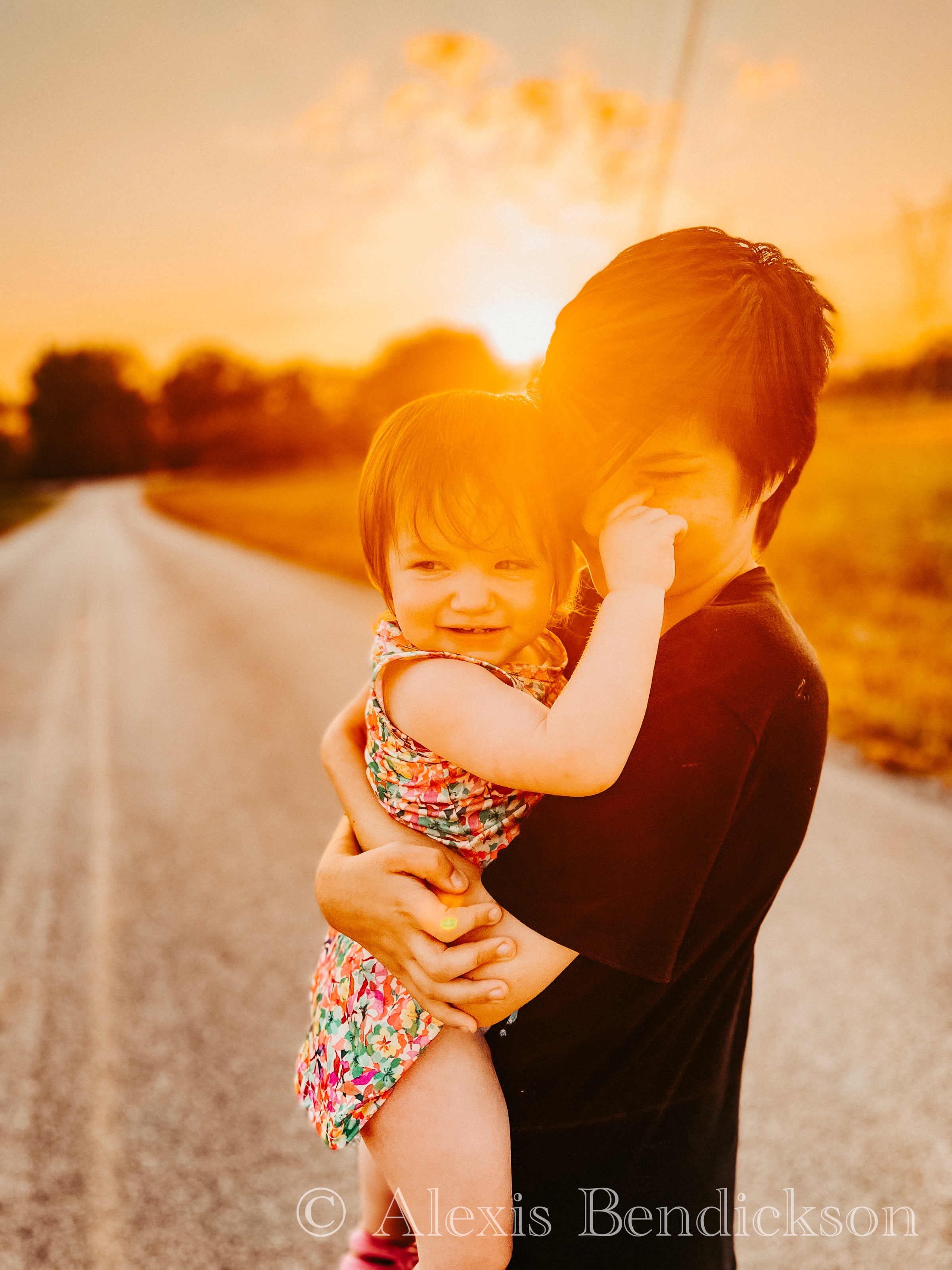 Emme smiling while being held by Noah at sunset, warm sun flare