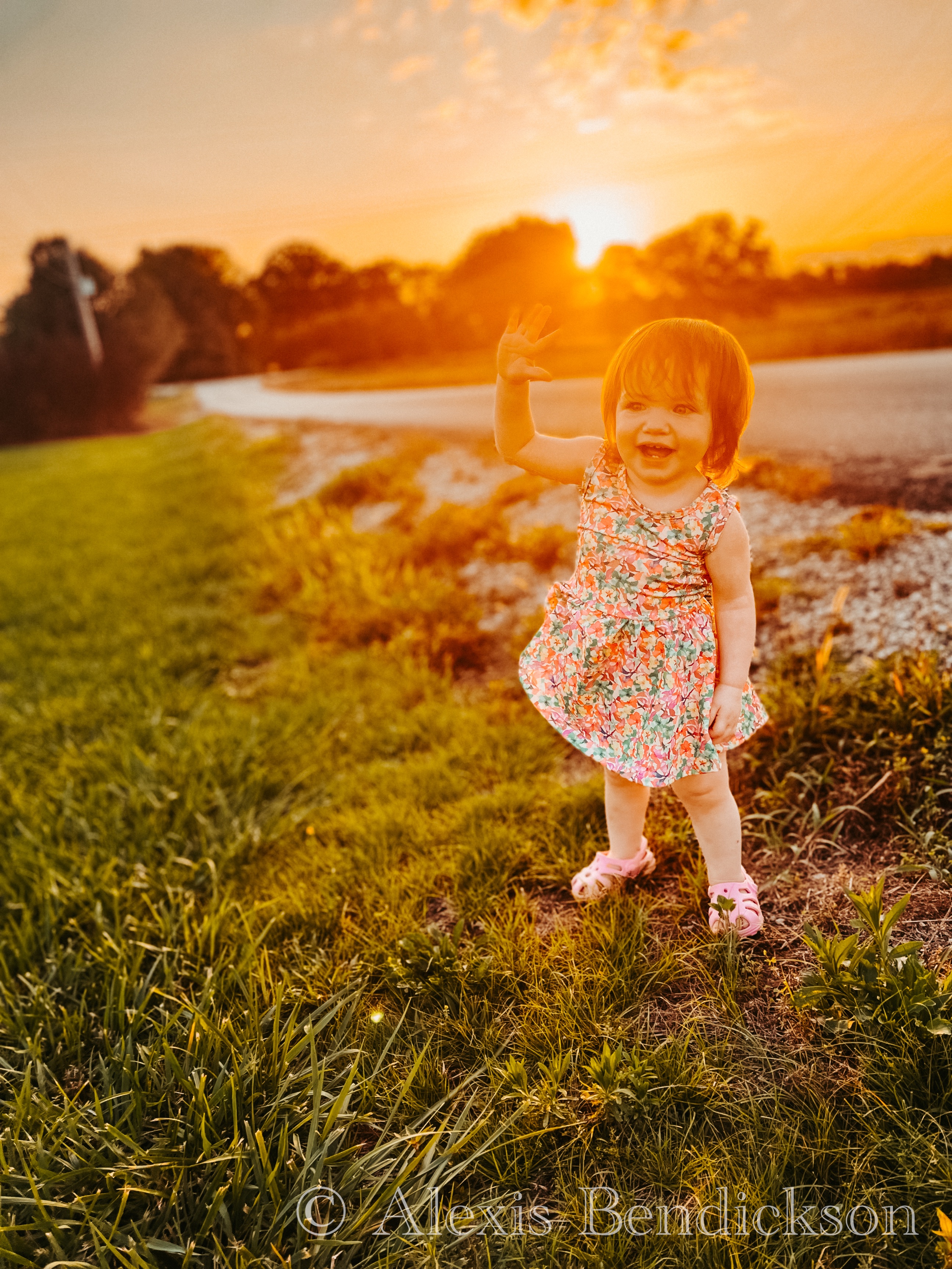Emme in a floral dress waving at the camera in a grassy field at sunset