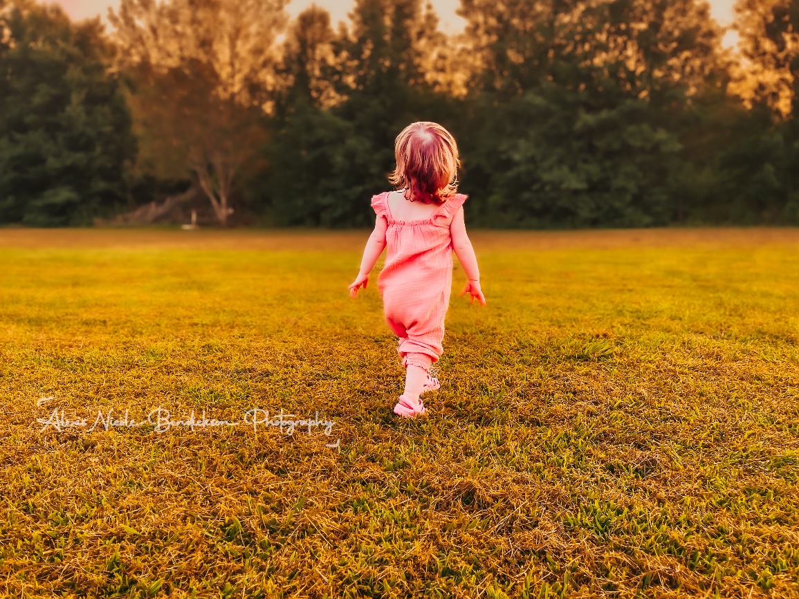 A toddler in a pink romper running across a field at sunset, seen from behind