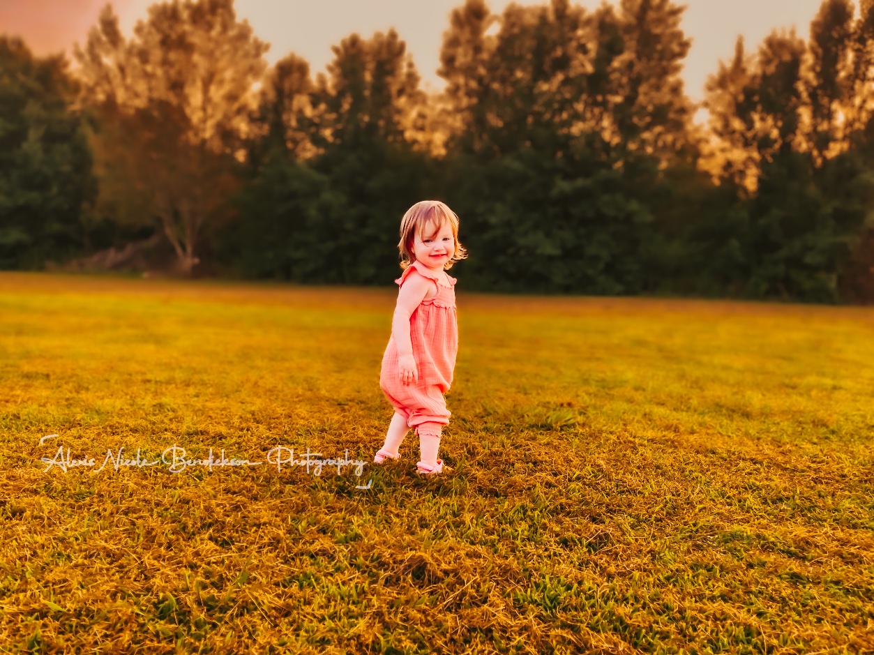 A toddler in a pink romper smiling back at the camera in a field at sunset