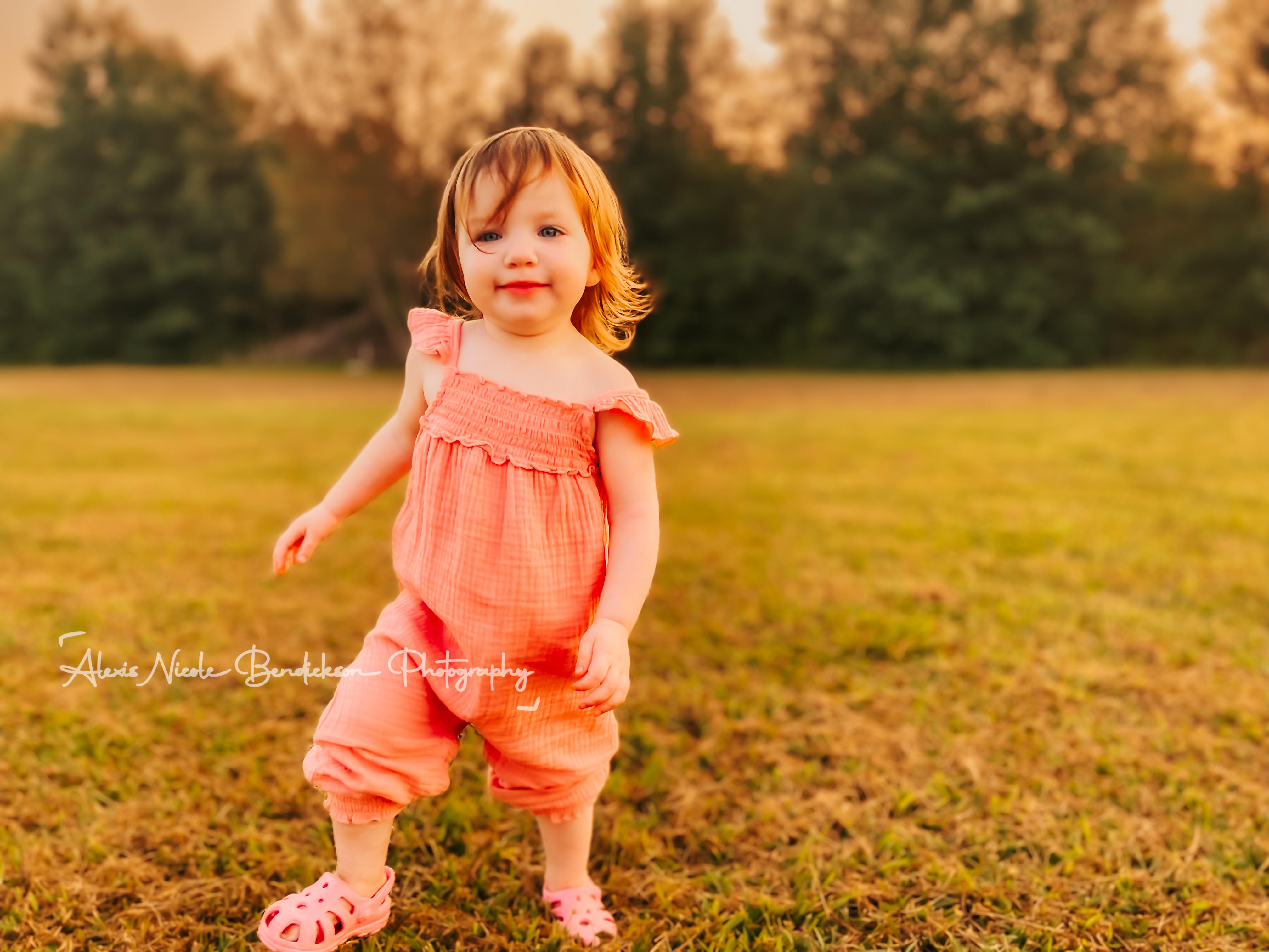 A toddler in a pink romper standing in a field at sunset