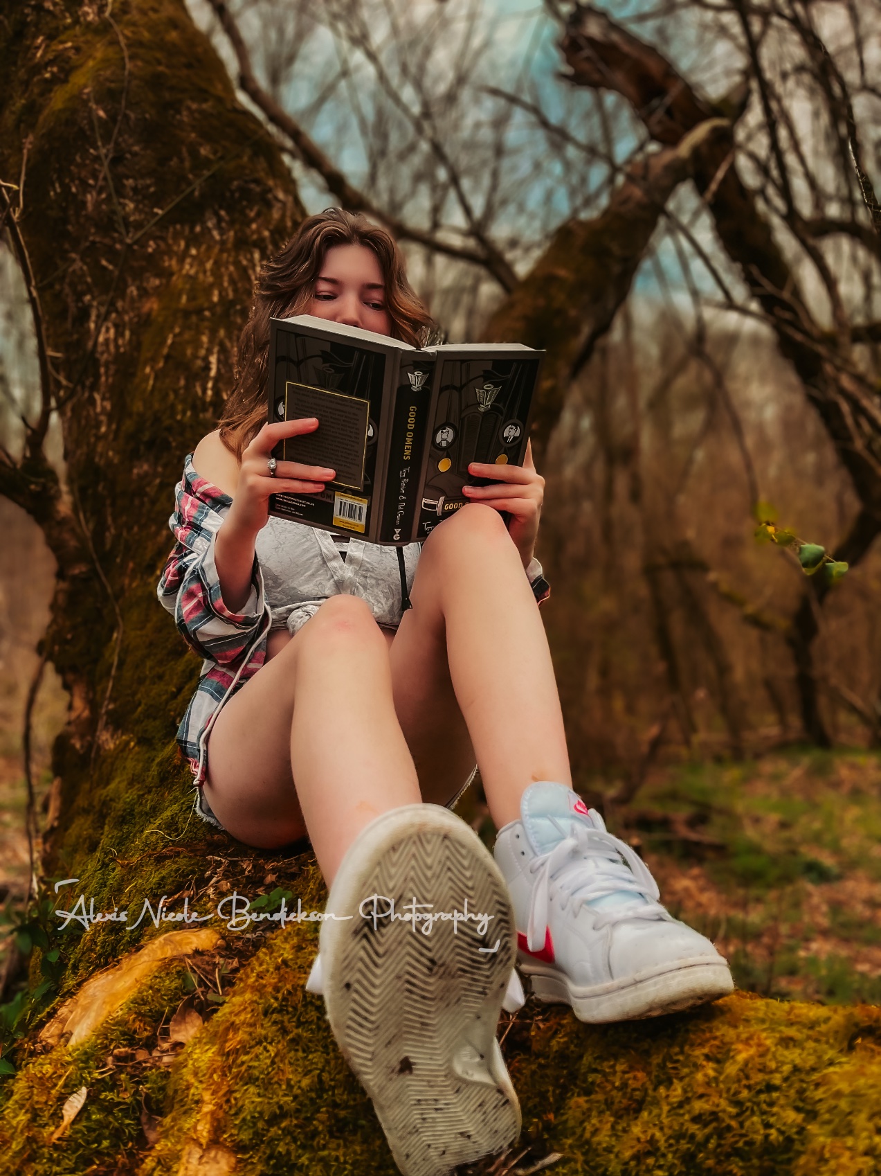 A young woman reading with the book held up, seated on a mossy tree