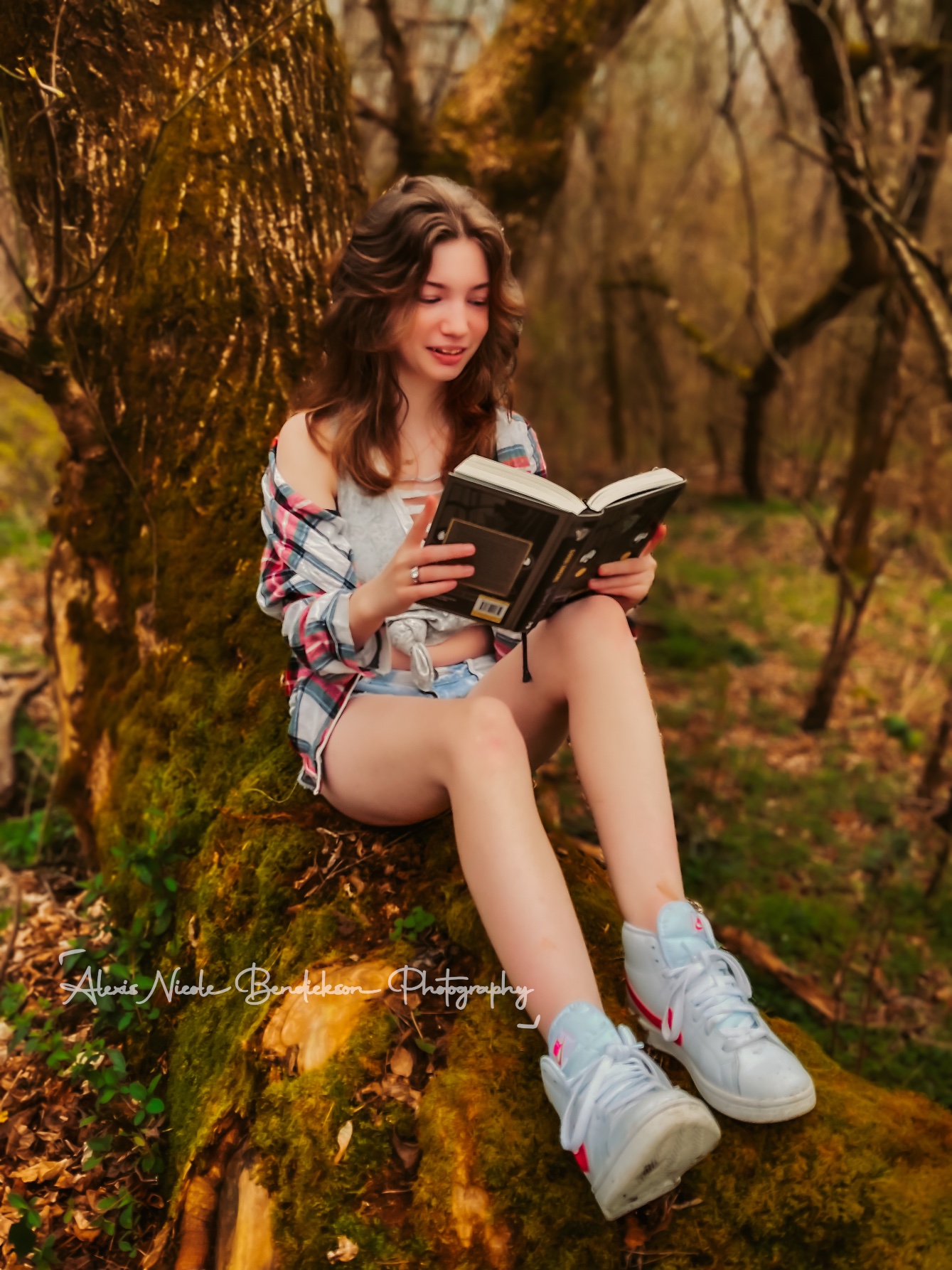 A young woman reading while seated on a moss-covered tree in the woods