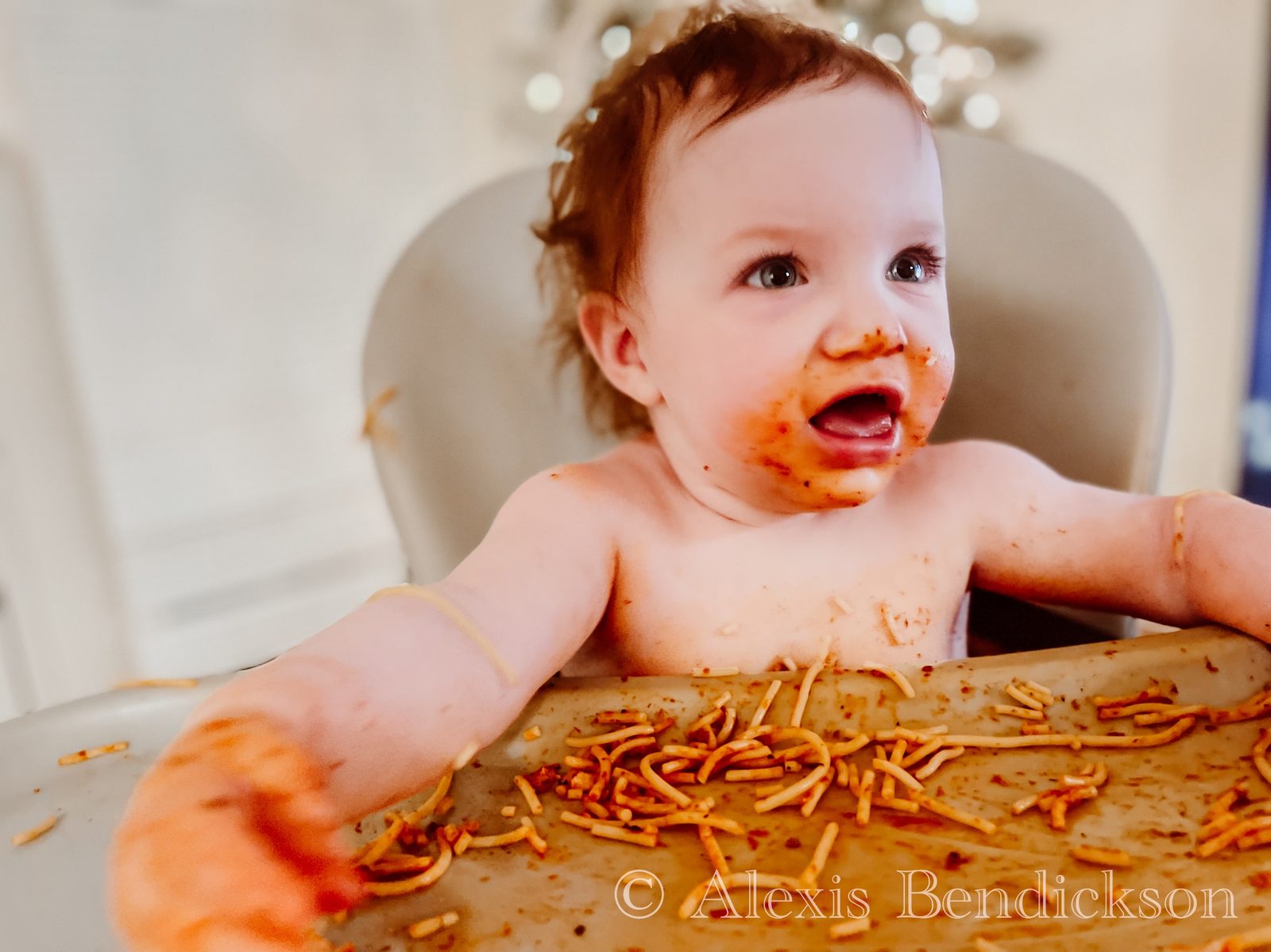 Baby Emme covered in spaghetti in her high chair, laughing