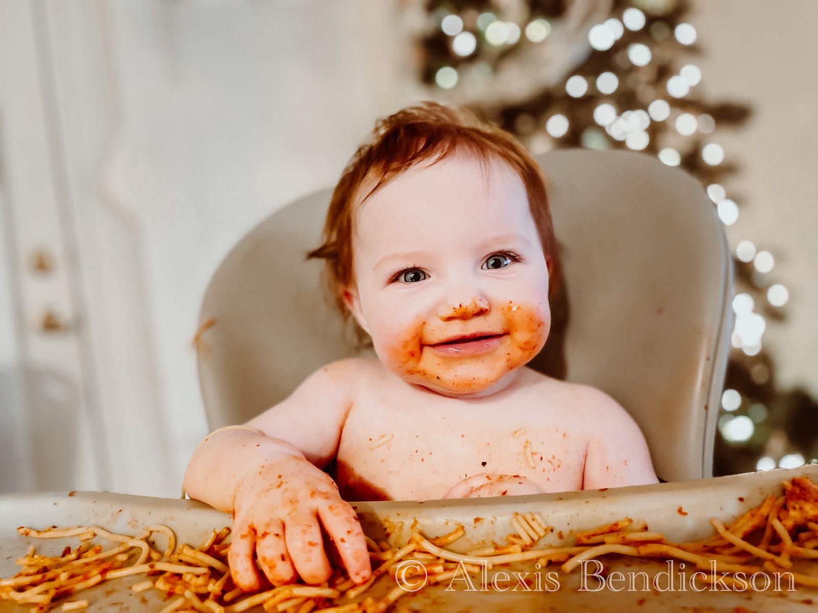 Baby Emme smiling with spaghetti on her face, Christmas lights behind her