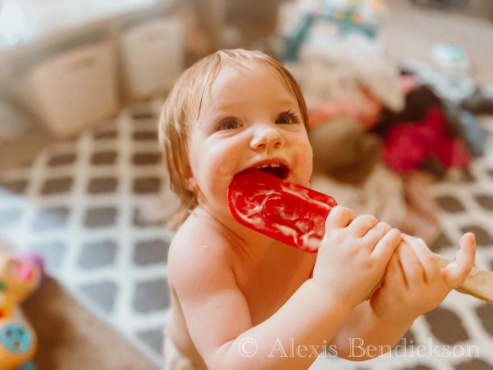 Toddler Emme grinning with a red spatula in her mouth