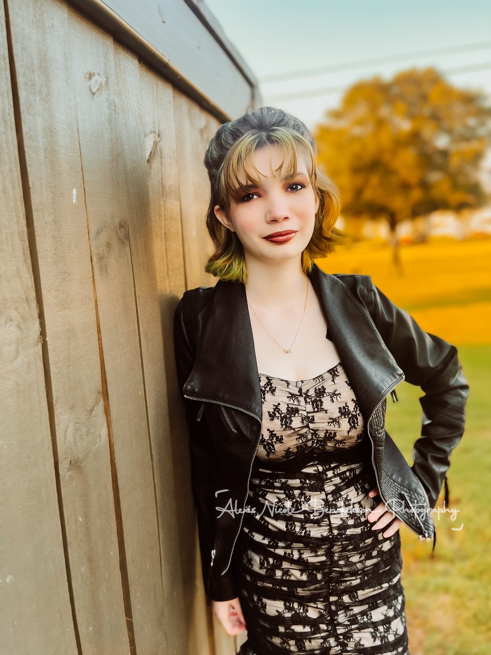 Lillia leaning against a wooden fence with golden autumn trees behind her