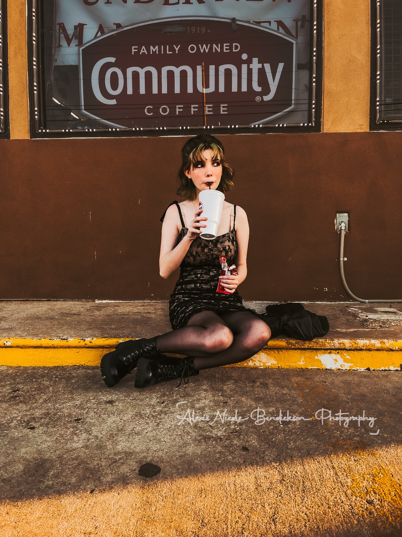 Lillia sitting on a curb drinking from a tall cup under a coffee shop window