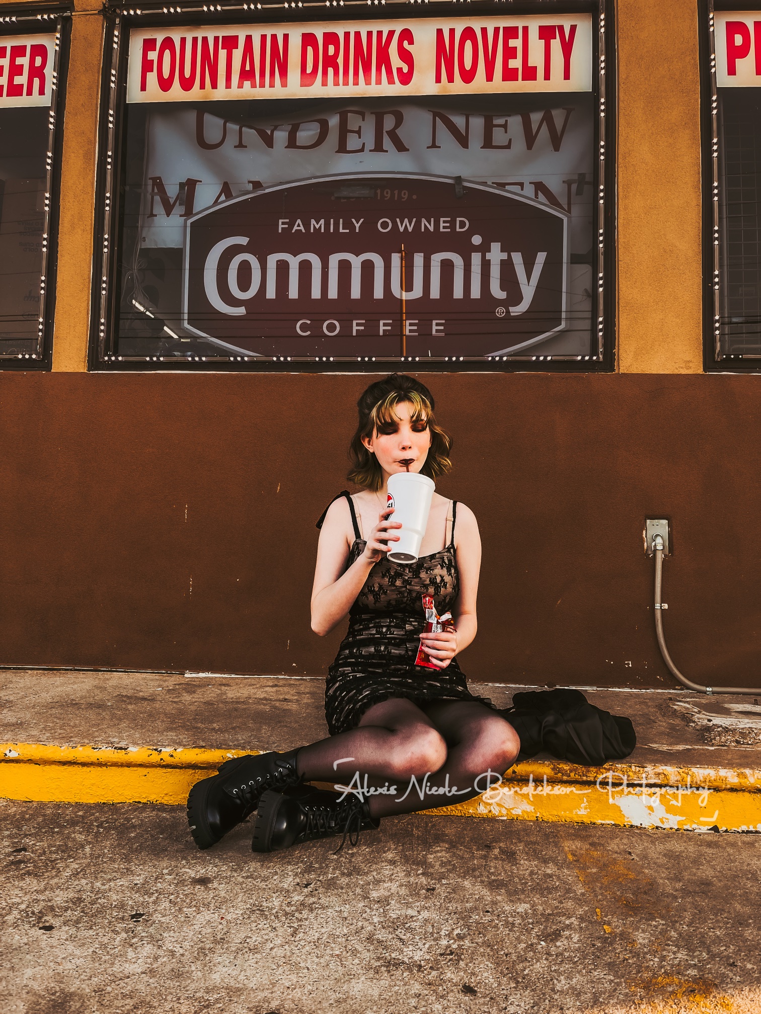 Lillia drinking from a tall cup beneath a 'fountain drinks novelty' sign
