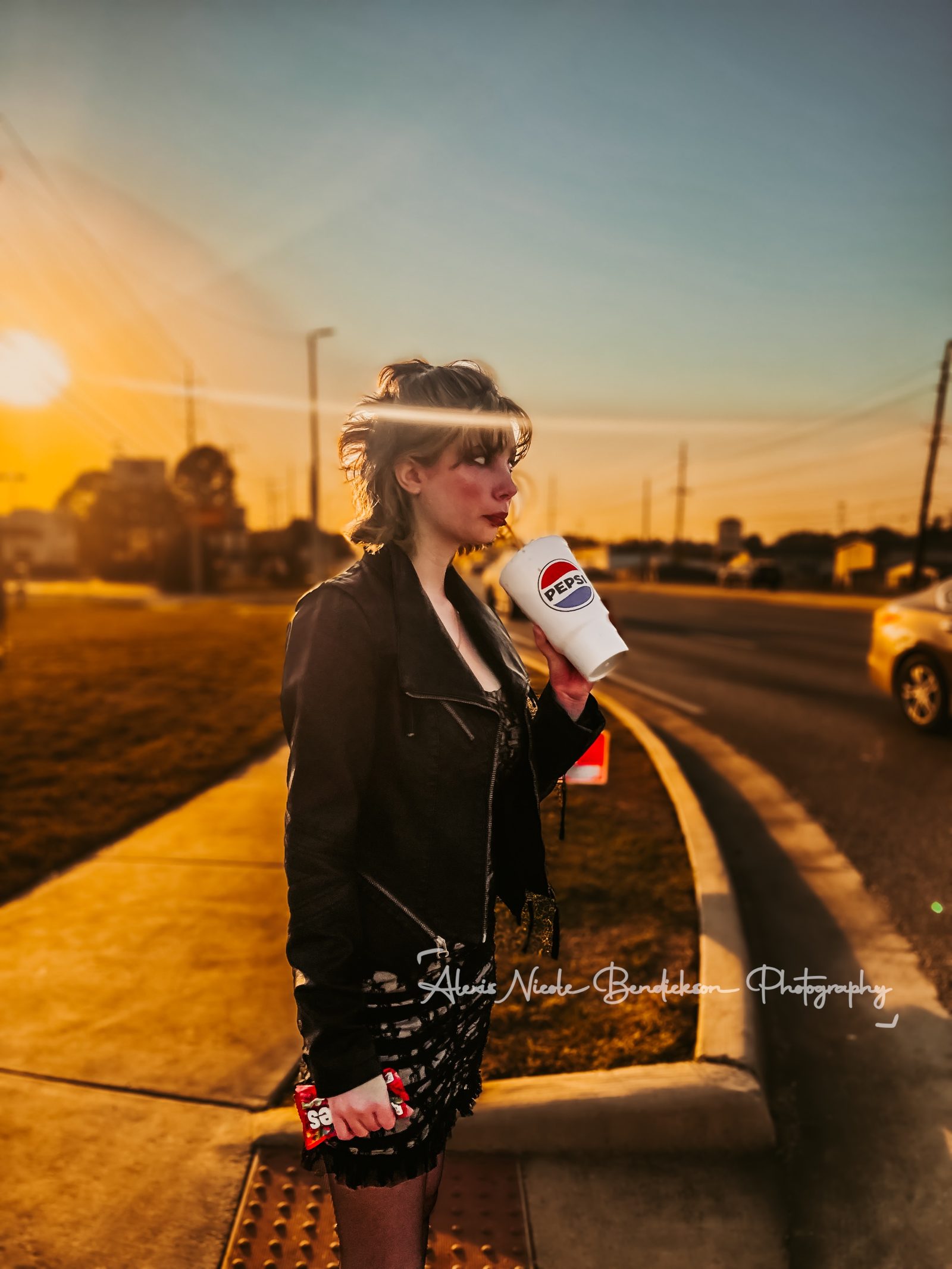 Lillia in a leather jacket drinking from a soda cup on a sidewalk at sunset