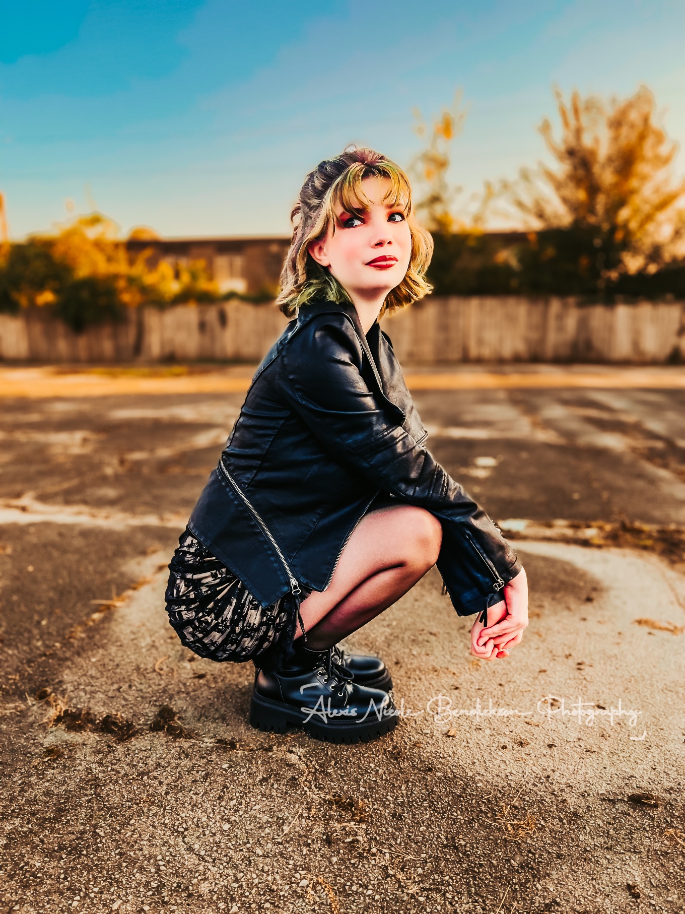 Lillia crouching in an empty parking lot in a leather jacket at golden hour