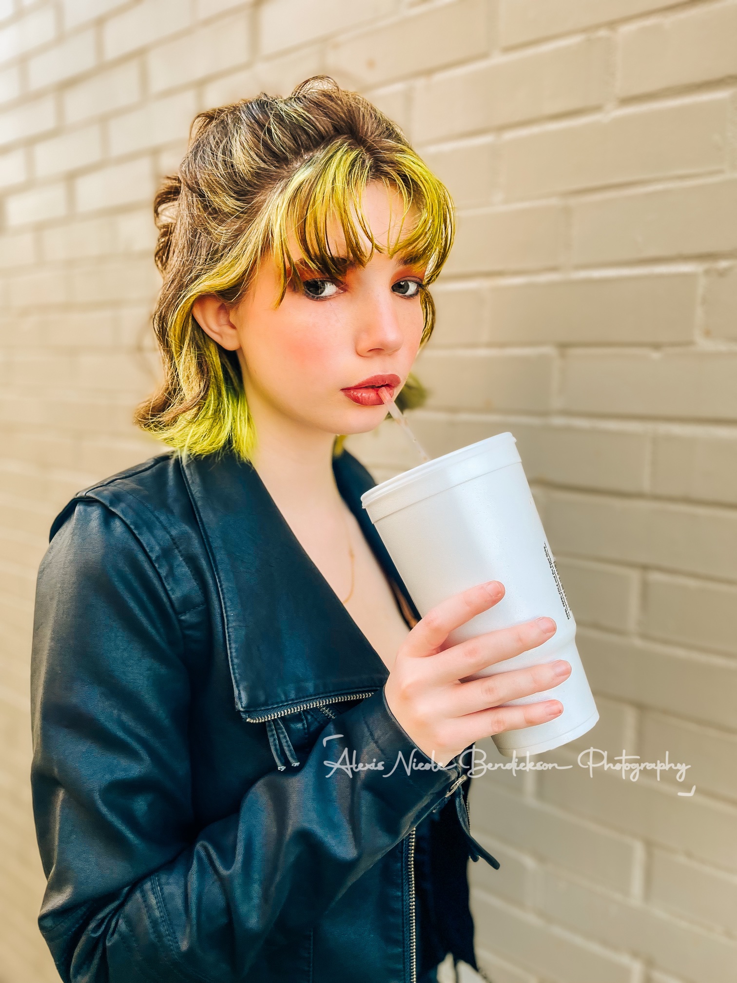 Lillia with a tall cup against a white brick wall